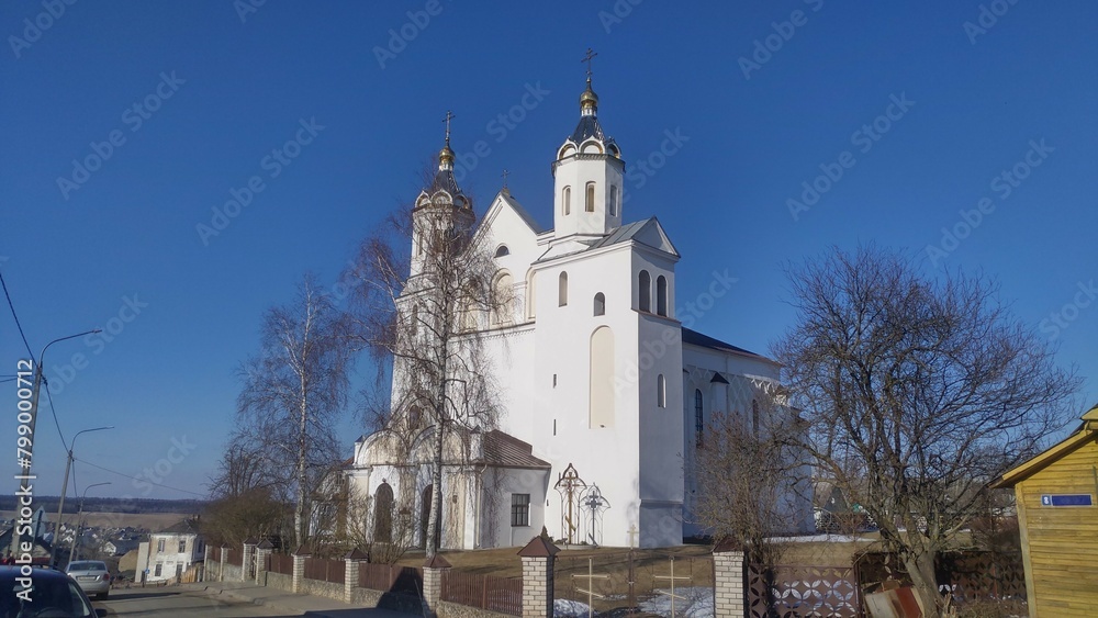 A white plastered Orthodox church with tall towers and crosses stands on a city street surrounded by buildings and trees. Cars and power lines stand nearby. Sunny spring weather with remnants of snow