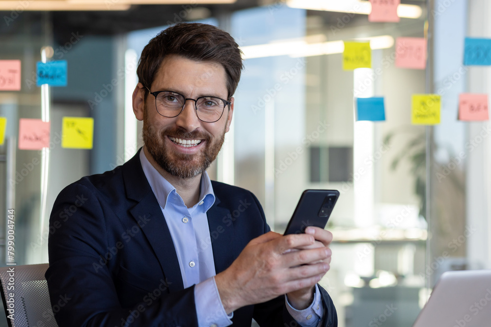 Portrait of successful man in business suit in office interior, mature businessman smiling and looking at camera, satisfied with achievement results, using app on phone. Portrait of successful man in business suit in office interior, mature businessman smiling and looking at camera, satisfied with achievement results, using app on phone.