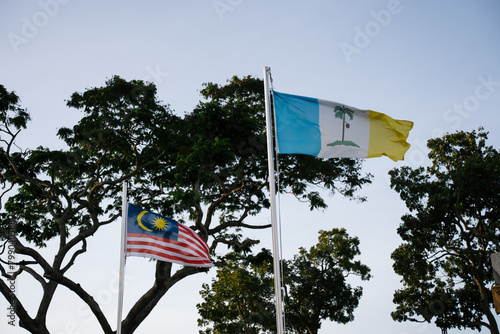 The Malaysia and Penang flags are waving in the wind against a background of trees and the sky.