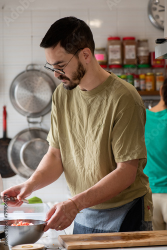 man preparing food, with a metal bowl, and woman in the background with her back to him, cooking together as a team. Two green T-shirts of different shades. Pans hanging on the wall