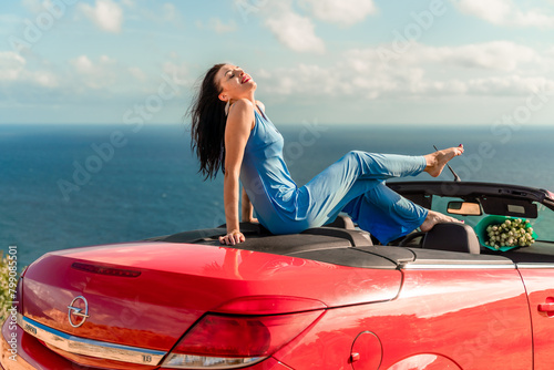 A woman is sitting in a red convertible car, smiling and enjoying the view of the ocean. Scene is happy and relaxed. © svetograph