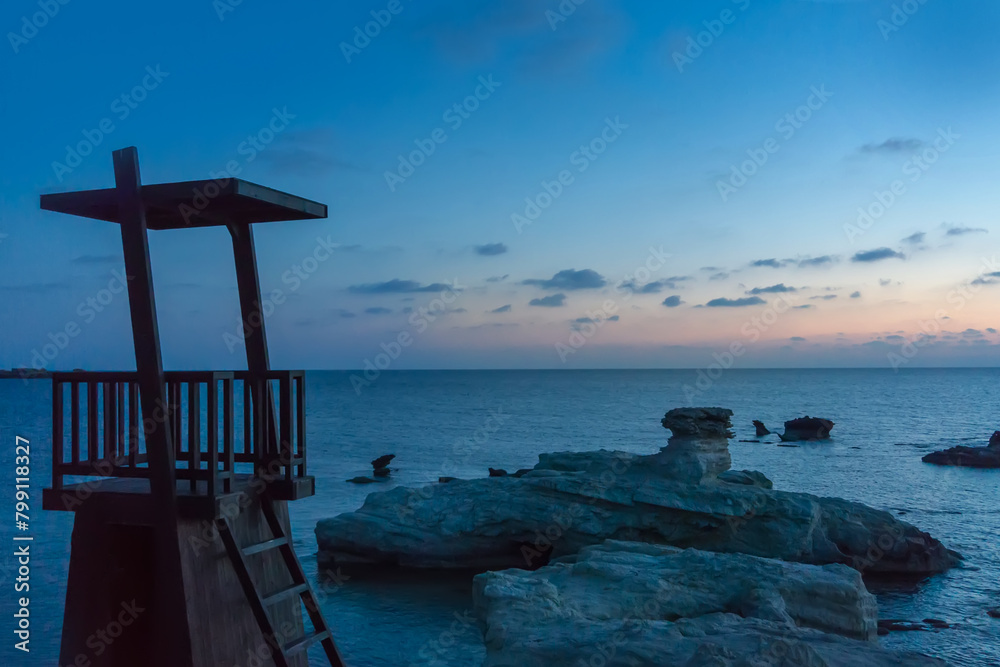 Serene Twilight Over a Coastal Lifeguard Tower at Dusk. In the foreground, a solitary lifeguard tower stands vigil atop rugged shoreline rocks, overlooking the calm sea Serene Twilight Over a Coastal Lifeguard Tower at Dusk. In the foreground, a solitary lifeguard tower stands vigil atop rugged shoreline rocks, overlooking the calm sea