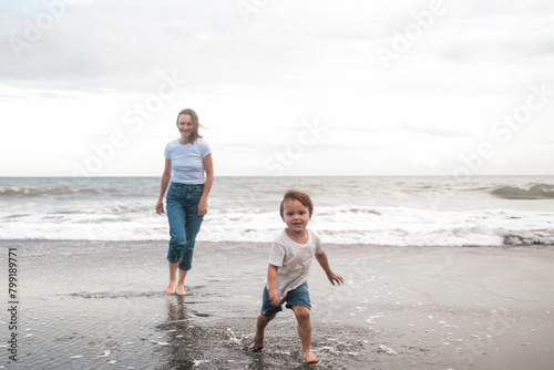 Mom and son Toddler are running along the black sand beach along the ocean and laughing happily. They play in the water and spend time together. Family summer vacation. Mothers Day. © moonmovie