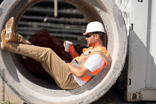 A construction worker at a construction site drinks coffee during a break. builder man in a helmet and vest holding a glass of coffee. a construction inspector in a helmet drinking coffee. photo