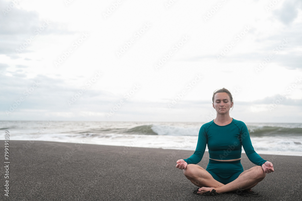 Banner with woman in lotus position sits on a black sand beach and meditates against the backdrop of the ocean with big waves. Practice mindfulness and purity of mind. Concentration. Summer sports.