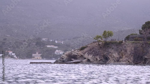 A rocky shoreline with a small town in the distance. The water is calm and the sky is overcast