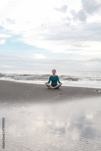 A young woman in lotus position sits on a black sand beach and meditates against the backdrop of the ocean with big waves. Practice mindfulness and purity of mind. Concentration. Summer sports.