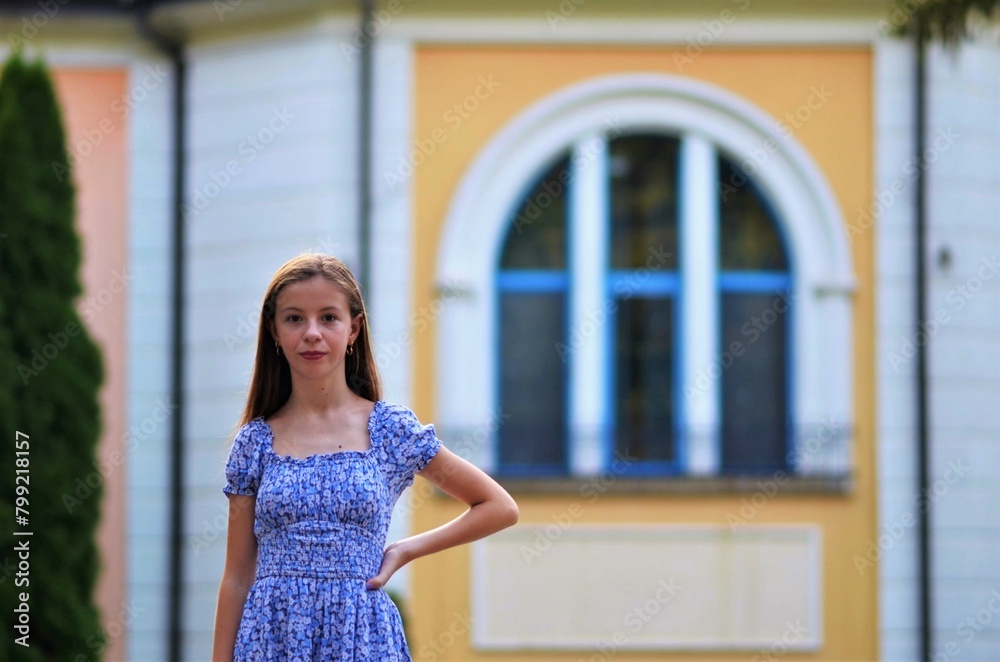 Fototapeta premium A young girl in a blue dress stands in front of a yellow building. The girl is smiling and looking at the camera. The scene is bright and cheerful, with the girl looking happy