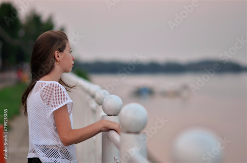 A girl is standing on a pier looking out at the water. The sky is pink and the water is calm