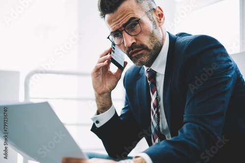 Confident successful businessman with financial text reports in hands calling to operator on smartphone working in office.Experienced proud ceo 50 years old with banking documents talking on telephone photo