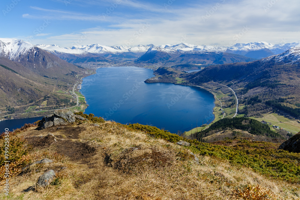 Overlooking a tranquil fjord, mountains rise in the distance beneath a bright, expansive sky.