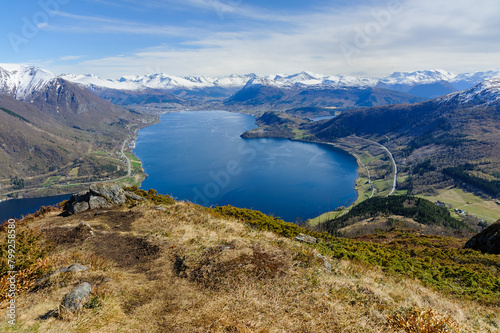 Overlooking a tranquil fjord, mountains rise in the distance beneath a bright, expansive sky.