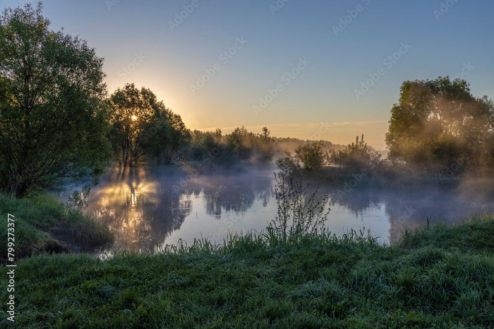 Fototapeta premium Blue fog over the river, the rising sun reflected in the water, foggy morning in the countryside.