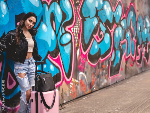 Urban stylish travel: young latina brunette with fashion look and pink suitcase in front of a lively street art mural at downtown