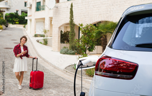 Woman traveler with a smartphone and bag next to charging electric car.. photo