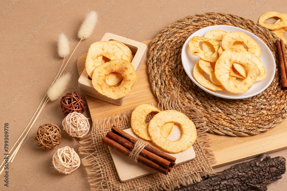 Crispy apple chips in a white round plate and in a wooden box, cinnamon sticks on a wooden desk on a beige background in rustic style. Healthy sweet snack. Crispy apple chips in a white round plate and in a wooden box, cinnamon sticks on a wooden desk on a beige background in rustic style. Healthy sweet snack.