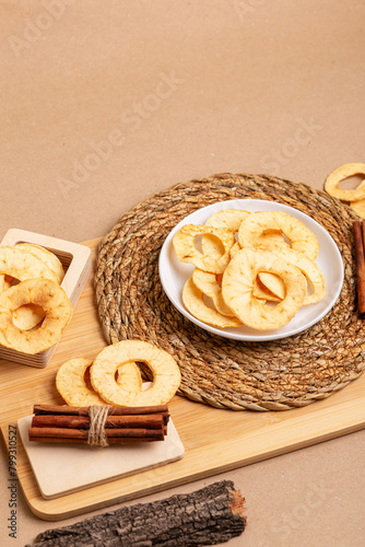 Crispy apple chips in a white round plate and in a wooden box, cinnamon sticks on a wooden desk on a beige background in rustic style. Healthy sweet snack.