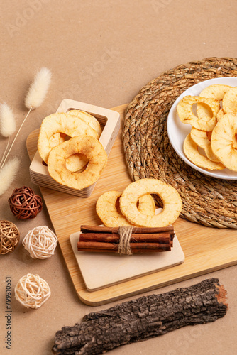 Crispy apple chips in a white round plate, cinnamon sticks on a wooden desk on a beige background in rustic style. Healthy sweet snack.