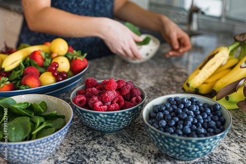 A close-up photograph of a woman arranging vibrant fruits and leafy greens on a kitchen counter, with bowls of ripe berries, bananas, and spinach ready to be blended into a nutriti