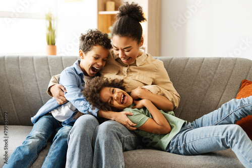 Happy two kids and their mother having fun while fooling around together, sitting on sofa at home