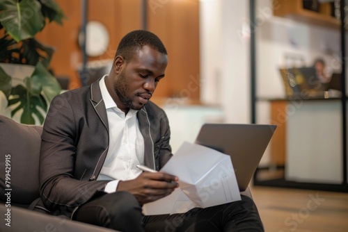 A businessman sits on a couch engaged in reviewing documents with a laptop, portraying concentrated work photo