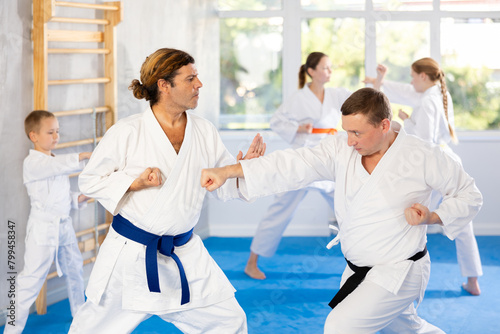 Man in kimono and colored belt practicing karate punch block whith trainer during group martial arts lesson in gym photo