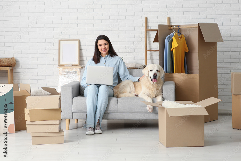 Fototapeta premium Young happy woman and cute Labrador dog with moving boxes sitting on sofa at home