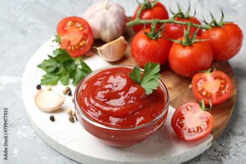 Delicious ketchup in bowl, parsley, garlic and tomatoes on grey textured table, closeup