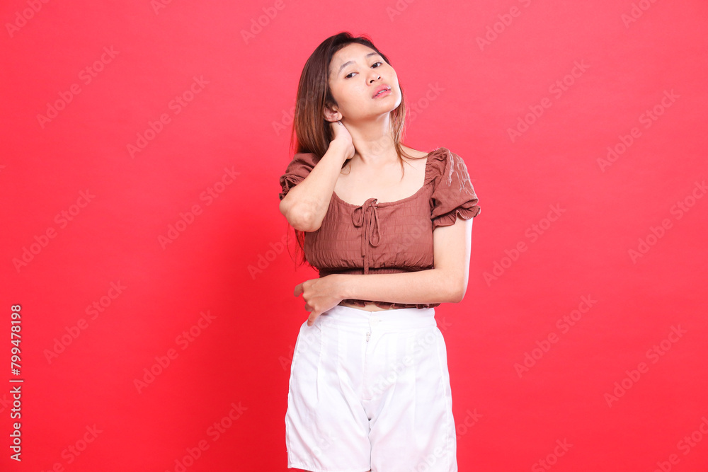 The expression of an Asian woman with her arms crossed, holding the back of her neck in pain, wearing a brown blouse and shorts with a red background. for health, care and advertising concept The expression of an Asian woman with her arms crossed, holding the back of her neck in pain, wearing a brown blouse and shorts with a red background. for health, care and advertising concept