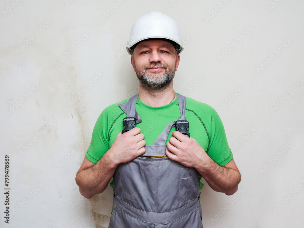 Portrait of a construction worker on light wall background. Builder in grey heavy duty clothes, white safety plastic helmet and green shirt. Male in his 40s with grey beard, athletic body type. Portrait of a construction worker on light wall background. Builder in grey heavy duty clothes, white safety plastic helmet and green shirt. Male in his 40s with grey beard, athletic body type.
