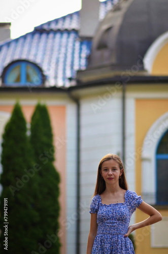 A young woman stands in front of a building with a blue dress on. She is smiling and looking at the camera
