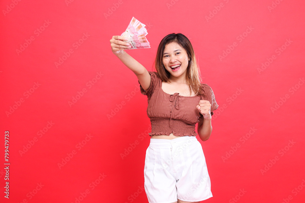 expression of a happy young Asian woman holding rupiah bills up and holding her hands to the side wearing a brown blouse top on a red background. for transaction, technology and advertising concepts expression of a happy young Asian woman holding rupiah bills up and holding her hands to the side wearing a brown blouse top on a red background. for transaction, technology and advertising concepts