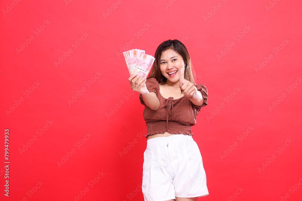 gesture of a cheerful young Asian woman holding rupiah money forward and giving a thumbs up sign (okay cool) wearing a brown blouse top with a red background. for transaction and advertising