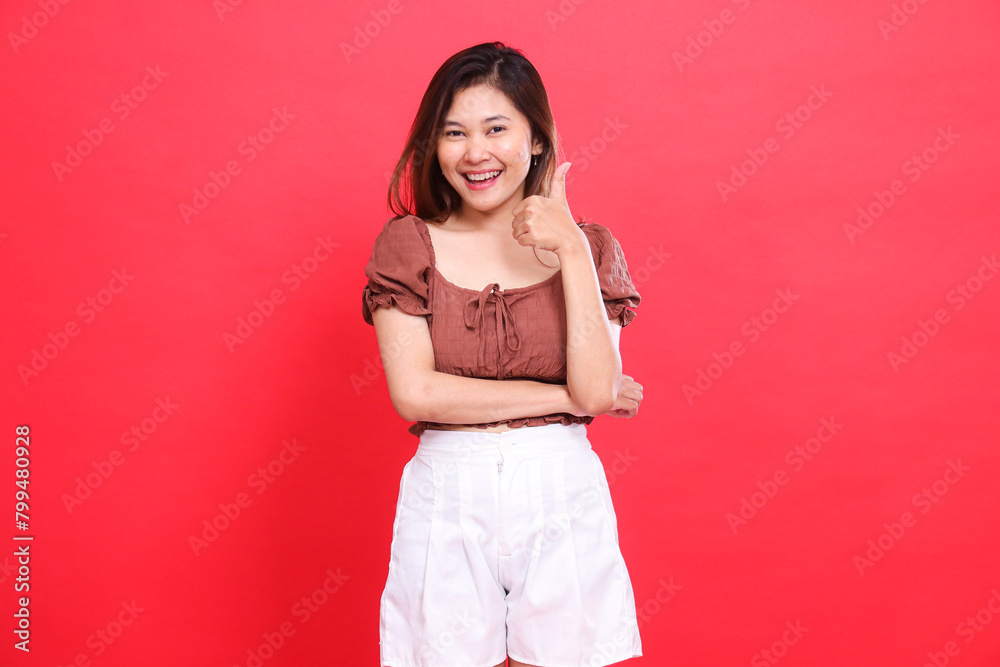 Cheerful indonesia woman's expression with her arms crossed with a thumbs up sign okay (cool) wearing a brown blouse and shorts with a red background. for fashion, lifestyle and advertising concepts Cheerful indonesia woman's expression with her arms crossed with a thumbs up sign okay (cool) wearing a brown blouse and shorts with a red background. for fashion, lifestyle and advertising concepts