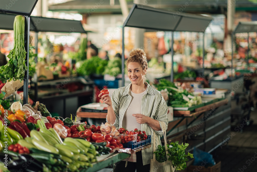 Happy woman at marketplace with tomato in hand smiling at camera.