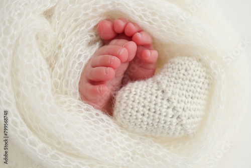 The tiny foot of a newborn baby. Soft feet of a new born in a white wool blanket. Close up of toes, heels and feet of a newborn. Knitted white heart in the legs of a baby. Macro photography