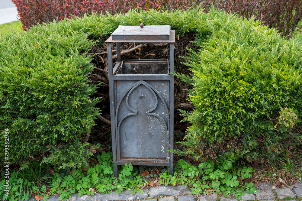 Gray metal trash can in ornamental green bushes. Gray metal trash can in ornamental green bushes.