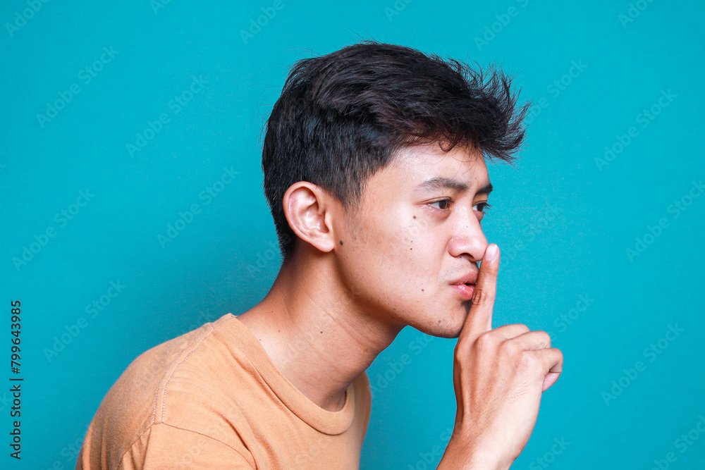 Side view of young Asian man keeps finger on lips, asks not tell secret information or keep silence Side view of young Asian man keeps finger on lips, asks not tell secret information or keep silence