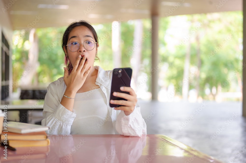 A shocked, excited Asian female college student covering her mouth while looking at her smartphone.