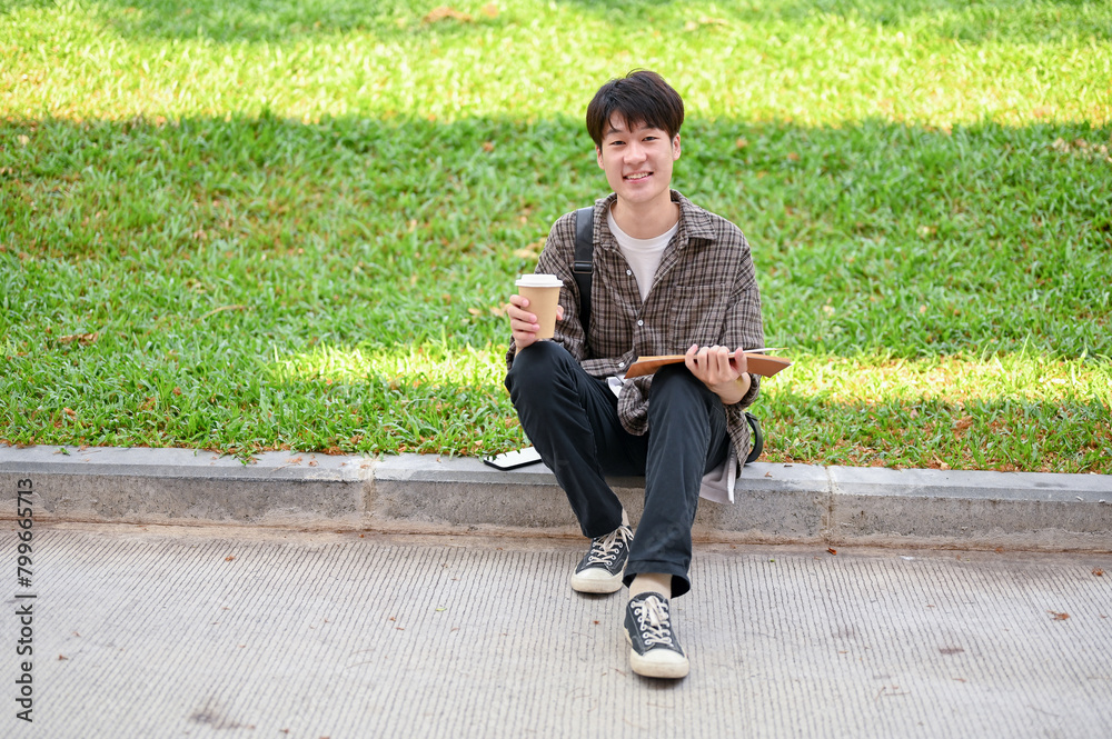 Obraz premium Smiling Asian student sits on the street in the campus park with a coffee cup and a book in his hand