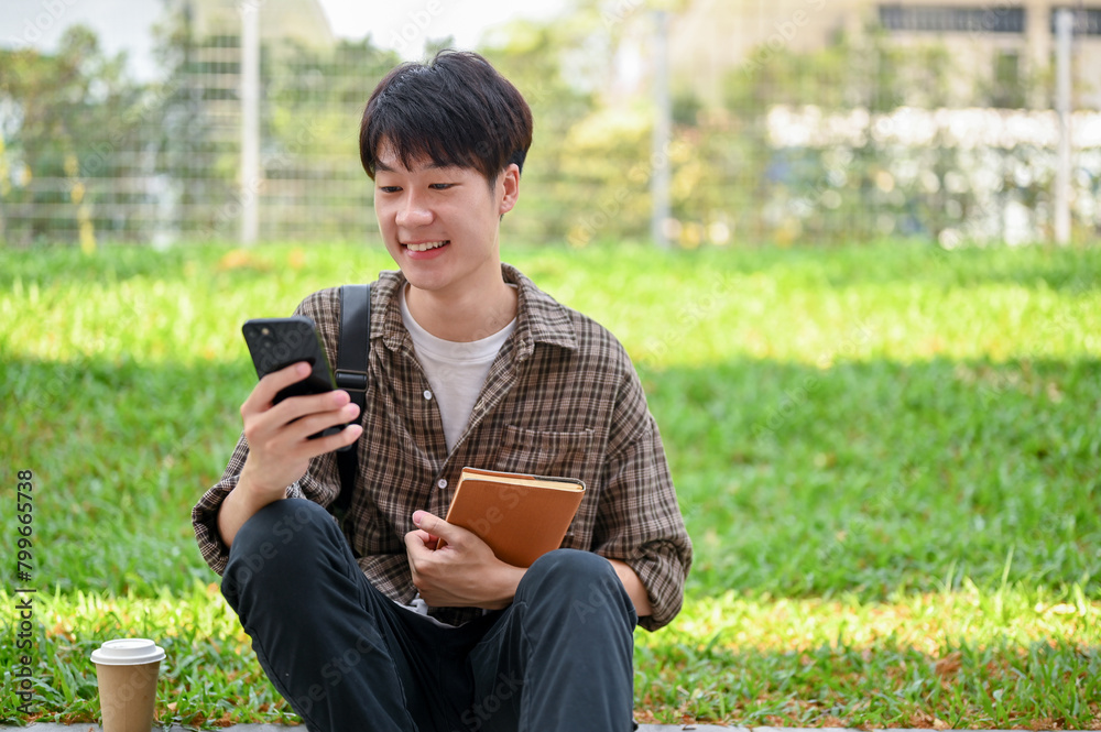 Obraz premium A cheerful Asian male college student sits on the street in the campus park using his phone.