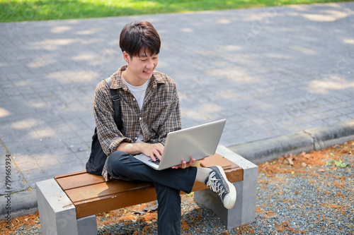 A young Asian male college student using his laptop computer on a bench in the campus park.