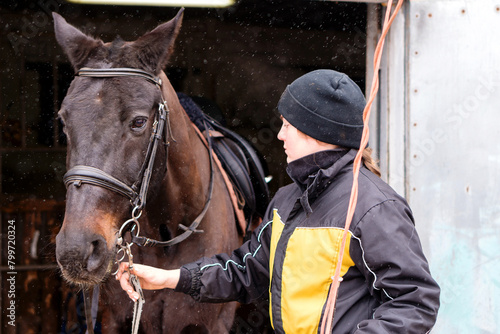 Person fastening horse's bridle in snowy barn.