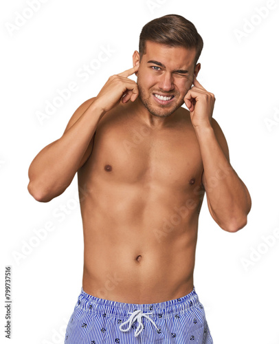 Young man shirtless in swimwear, studio covering ears with hands.