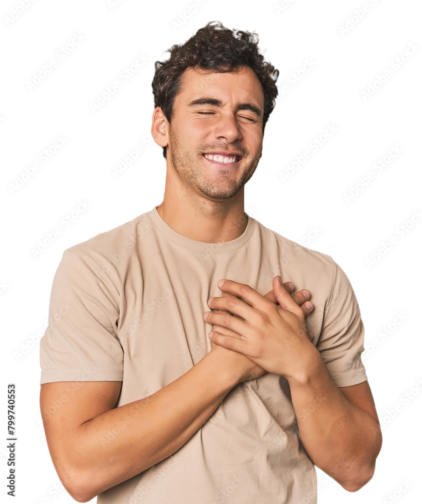 Young Hispanic man in studio laughing keeping hands on heart, concept of happiness.