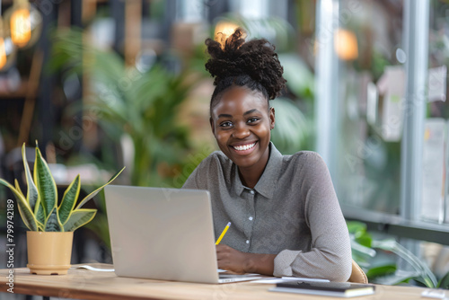 Photo of Smiling young african american woman writing on notepad while sitting at desk with laptop in office, real photo


