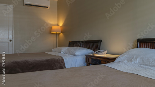 A fragment of the bedroom. Two beds are made with white sheets and brown bedspreads. The pillows are placed in the headboard. There's a phone on the bedside table. The floor lamp is lit in the corner