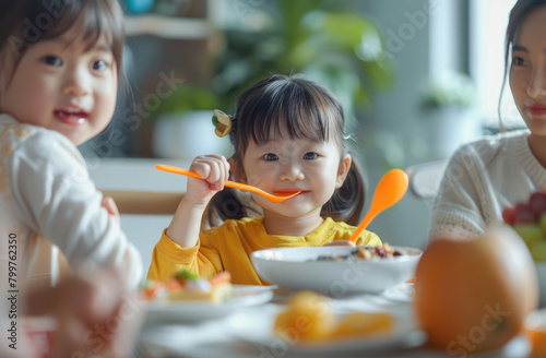 A young Asian girl is sitting at the dining table, holding an orange spoon in her hand and eating breakfast with family members around her. 