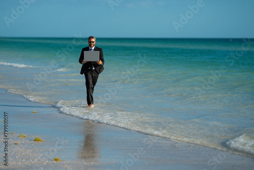Freelance work, online business. Summer business. Businessman in suit hold laptop and run on sea. Funny business man in a business suit with laptop standing in water. Remote work. Crazy freelancer.