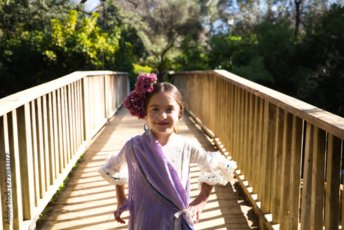 Portrait of a pretty girl dancing flamenco in a dress with frills and fringes typical of gypsies, walking on a wooden bridge in a famous park in Seville, Spain. Her hair is tied back with a flower.
